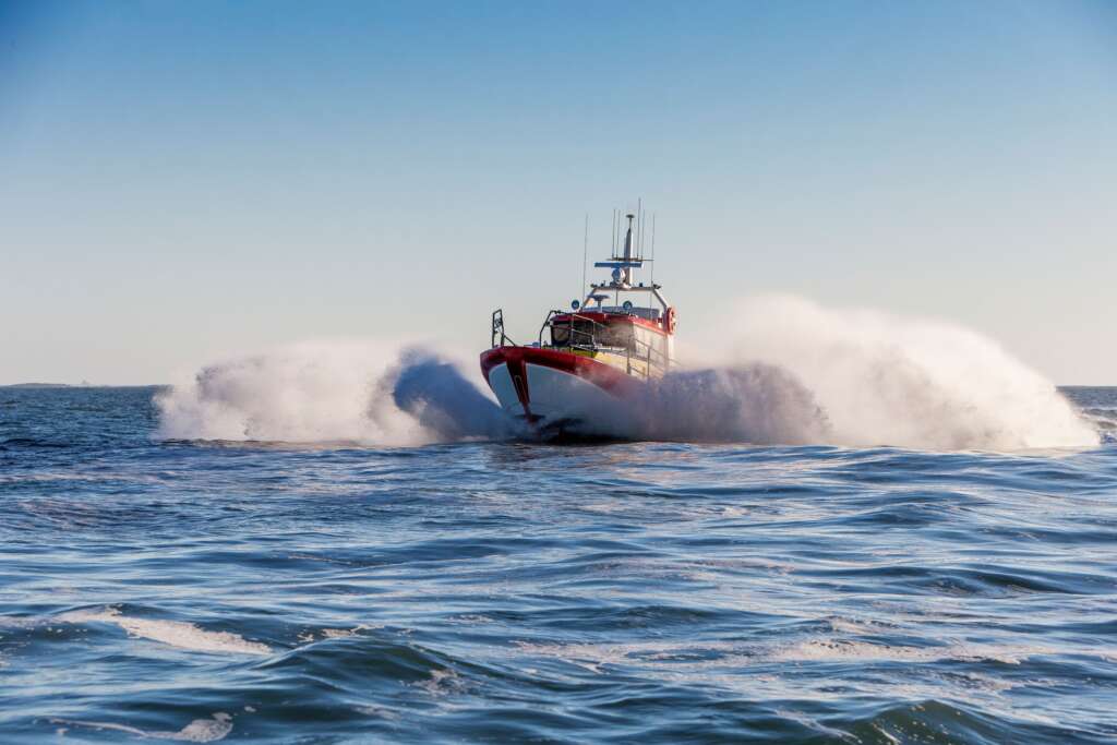 A Swedish Sea Rescue Society rescue boat travelling at speed across open water during an operational response, with waves and sea spray surrounding the vessel